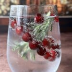 A clear glass of sparkling water with floating cranberries and a sprig of rosemary, set on a brown surface with a blurred, warm-toned background—an elegant twist inspired by the Viral Snow Globe Cocktail.