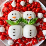 A plate of cherry tomatoes, mozzarella balls, and herbs, arranged with two Burrata Snowman figures made of mozzarella, decorated with peppercorn buttons, carrot noses, and basil leaf scarves.