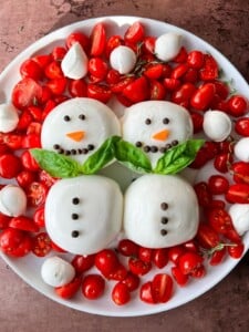 A plate of cherry tomatoes, mozzarella balls, and herbs, arranged with two Burrata Snowman figures made of mozzarella, decorated with peppercorn buttons, carrot noses, and basil leaf scarves.