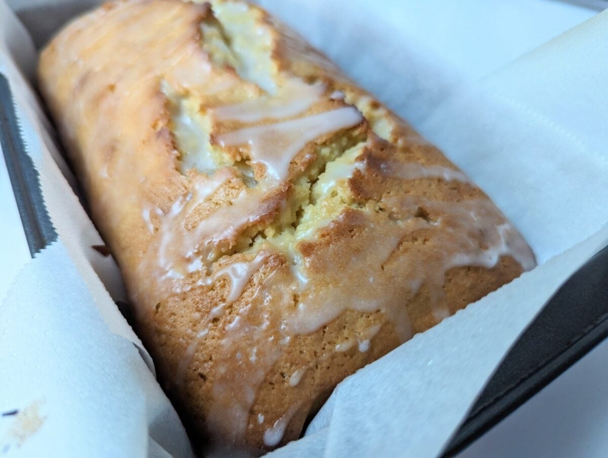 A loaf of Whole Lemon Blender Cake sits in a baking pan lined with parchment paper. The cake has a golden-brown crust with visible cracks and a light drizzle of icing on top.