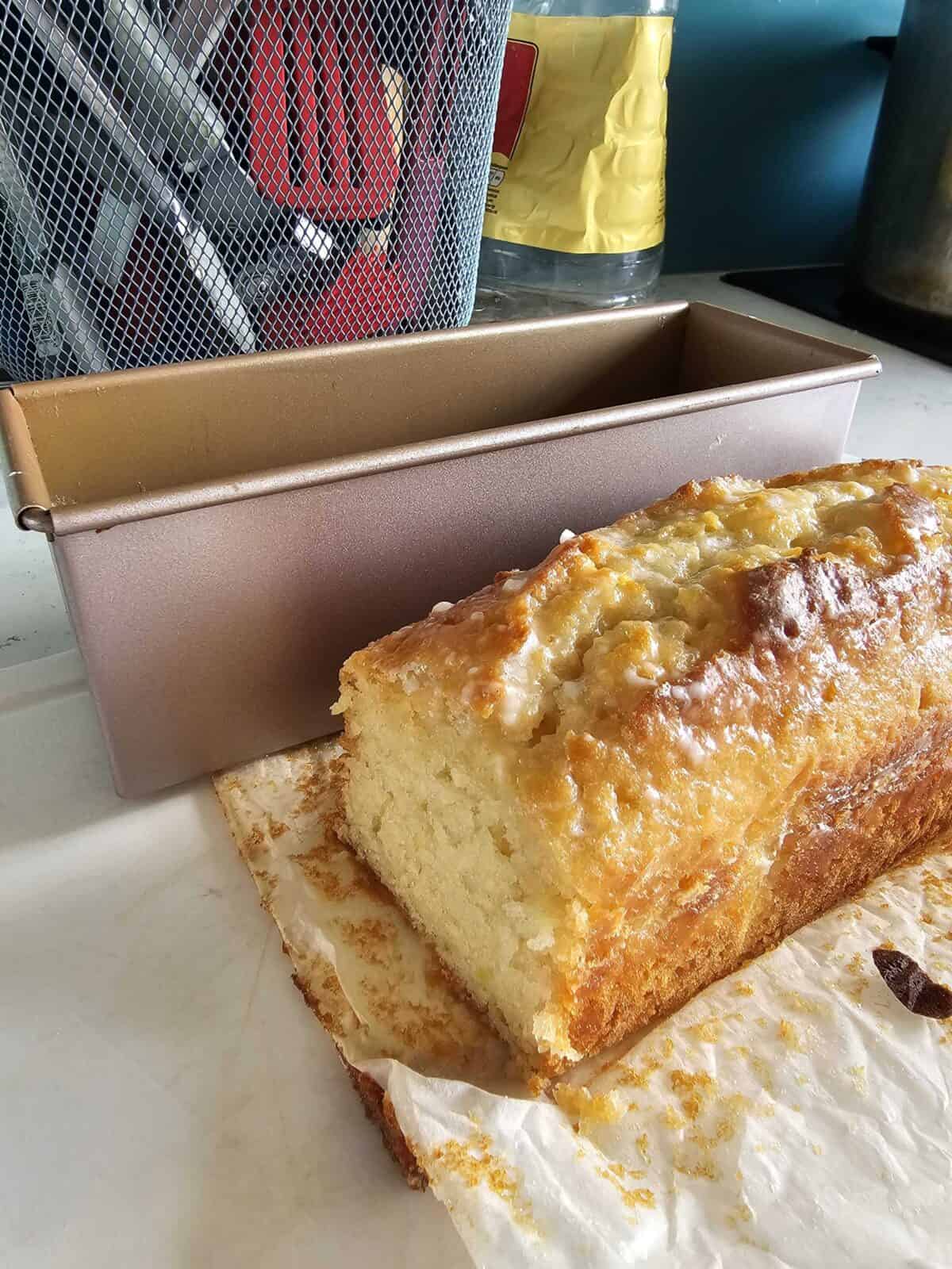 A loaf of golden, homemade bread sits on parchment paper beside a baking pan, with kitchen utensils and a bottle in the background. The bread appears moist with a slightly crisp crust.