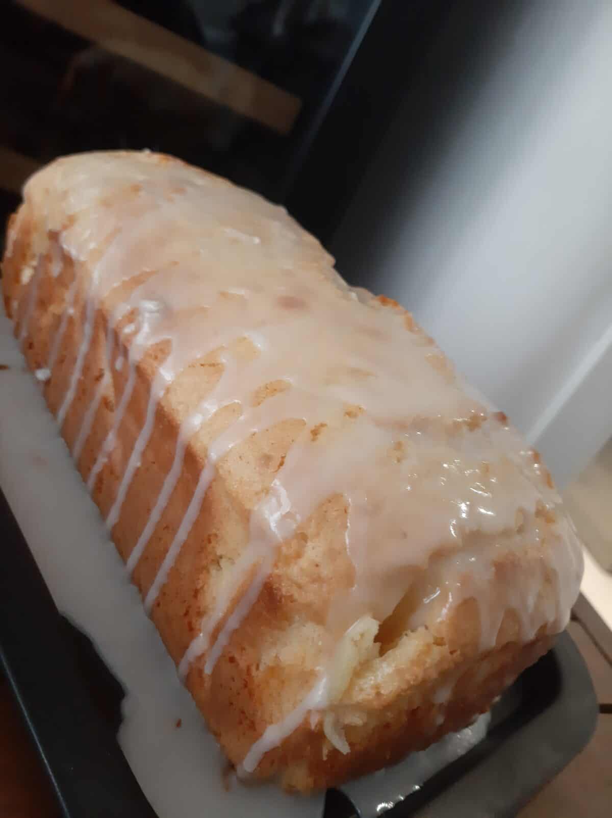 A Whole Lemon Blender Cake loaf with a golden crust is covered in a glossy white glaze that drips down the sides, sitting on a dark rectangular tray.