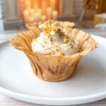 A Healthy Carrot Cake Muffin in a brown paper liner topped with creamy frosting and chopped nuts sits on a white plate. In the softly blurred background, lit candles and wooden decor add to the cozy atmosphere.