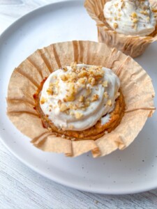 A Healthy Carrot Cake Muffin with white frosting and chopped nuts sits in a brown paper liner on a white plate, with another similar muffin blurred in the background.