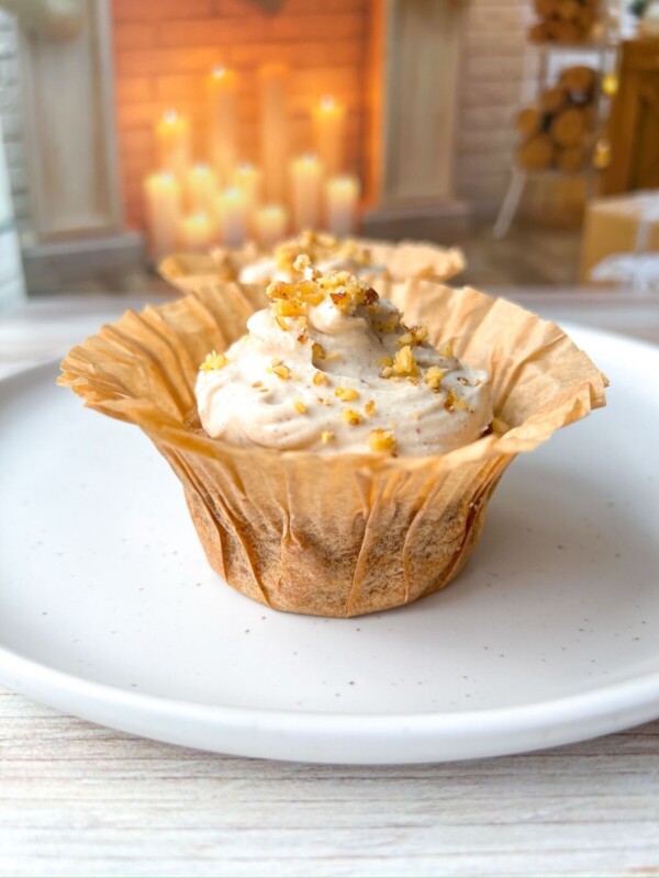 A Healthy Carrot Cake Muffin in a brown paper liner topped with creamy frosting and chopped nuts sits on a white plate. In the softly blurred background, lit candles and wooden decor add to the cozy atmosphere.