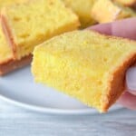 A hand holding a slice of moist yellow Clementine Cake with more slices stacked on a white plate in the background on a light wooden surface.