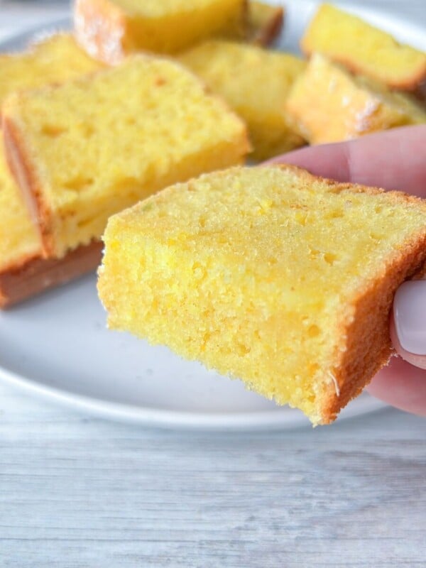 A hand holding a slice of moist yellow Clementine Cake with more slices stacked on a white plate in the background on a light wooden surface.