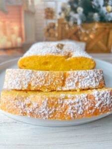 A close-up of a sliced clementine cake dusted with powdered sugar on a white plate, with a blurred holiday background featuring candles, wood, and a decorated Christmas tree.
