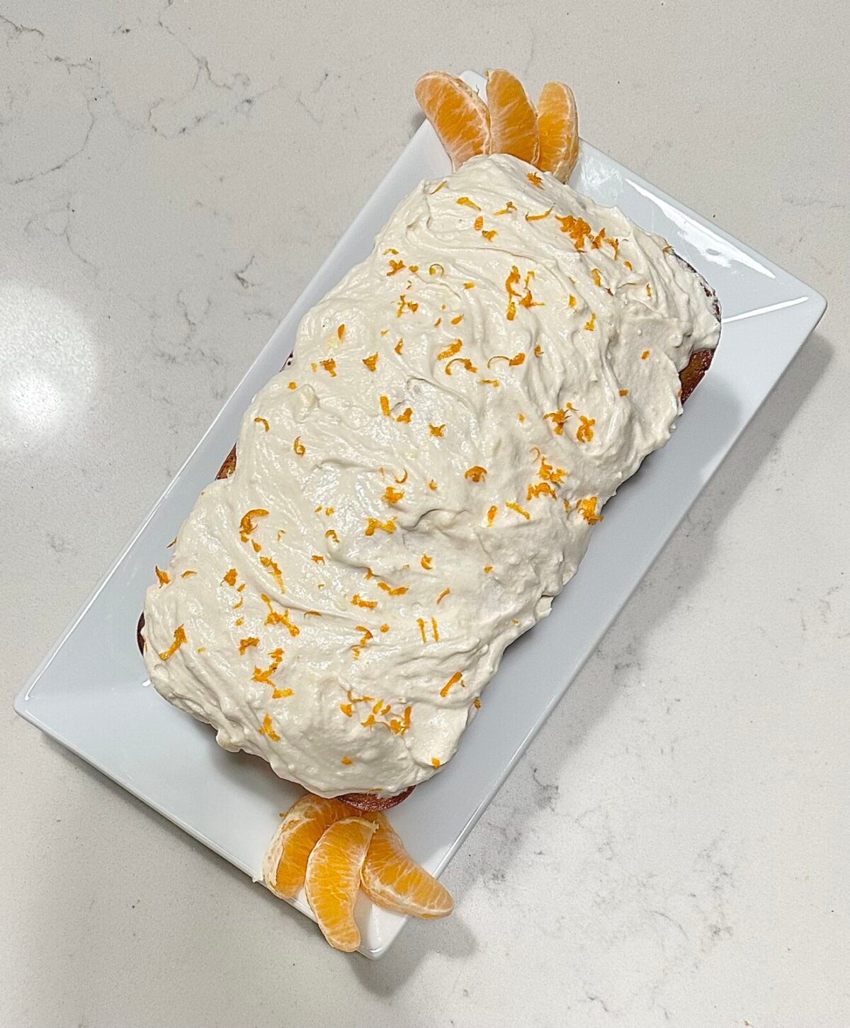 A Clementine Cake topped with swirled white frosting and orange zest sits on a rectangular white plate. Orange segments are placed at each end for decoration, while the plate rests on a light-colored marble countertop.