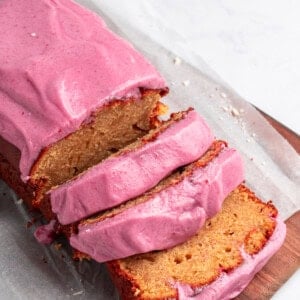 Loaf of cake with bright pink frosting, partially sliced, on a wooden board. White chocolate shavings scattered in the background on a light surface.