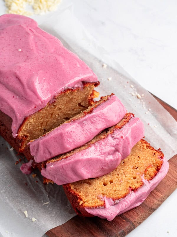 Loaf of cake with bright pink frosting, partially sliced, on a wooden board. White chocolate shavings scattered in the background on a light surface.