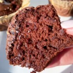 A close-up of a hand holding a half-eaten Healthy Chocolate Muffin, showing its moist and fluffy texture. Two more Healthy Chocolate Muffins in golden wrappers are visible in the background on a white plate.