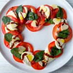 Heart Shaped Caprese Salad featuring sliced tomatoes, mozzarella, mint leaves, and heart-shaped mozzarella pieces arranged in a heart on a white plate, drizzled with balsamic glaze and olive oil.