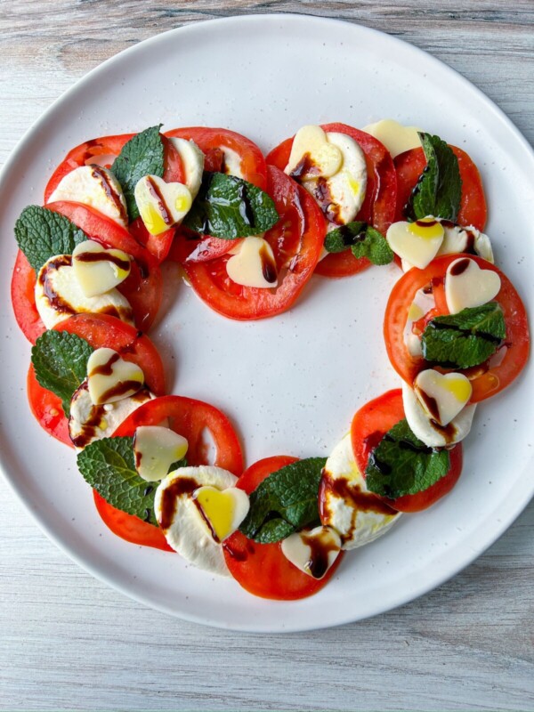 Heart Shaped Caprese Salad featuring sliced tomatoes, mozzarella, mint leaves, and heart-shaped mozzarella pieces arranged in a heart on a white plate, drizzled with balsamic glaze and olive oil.