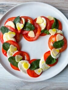A Heart Shaped Caprese Salad features sliced tomatoes, mozzarella, mint leaves, and small heart-shaped cheese pieces on a white plate, finished with drizzles of olive oil.
