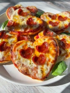 A plate of Heart Shaped Pizza minis, topped with melted cheese and small pepperoni slices, garnished with fresh basil leaves, sits in bright natural sunlight.