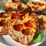 A plate of delightful Heart Shaped Pizza minis topped with melted cheese, small pepperoni slices, and fresh basil leaves sits on a white plate, basking in bright sunlight.