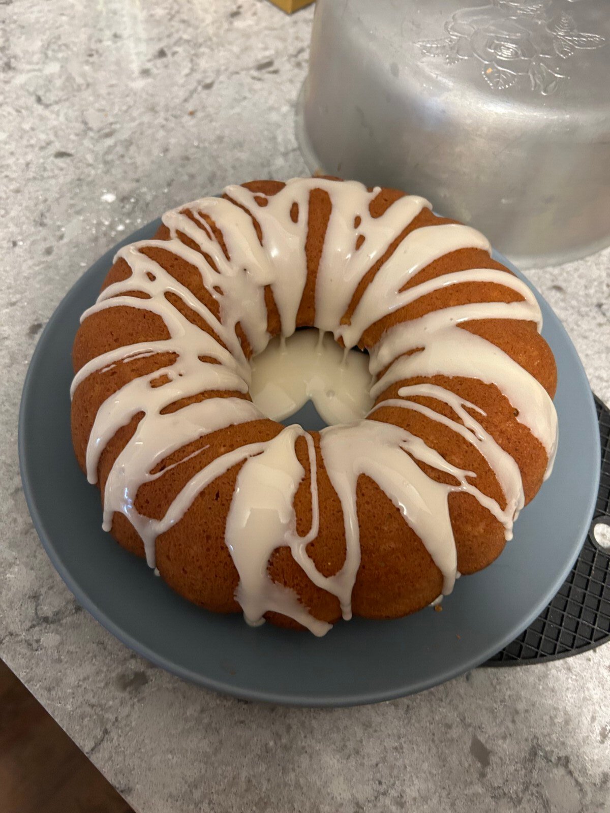 A round Clementine Cake with white icing drizzled on top sits on a blue plate on a marble countertop. A metal cake cover is partially visible in the background.
