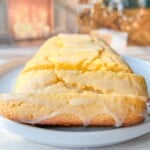 A close-up of a glazed Whole Lemon Blender Cake on a white plate. The cake has shiny lemon icing drizzling down the sides. In the blurred background, there are holiday decorations and a lit fireplace.