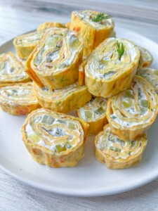 A plate of Zucchini Egg Roll pinwheels filled with cream cheese and diced vegetables, garnished with a sprig of fresh dill, displayed on a white plate.