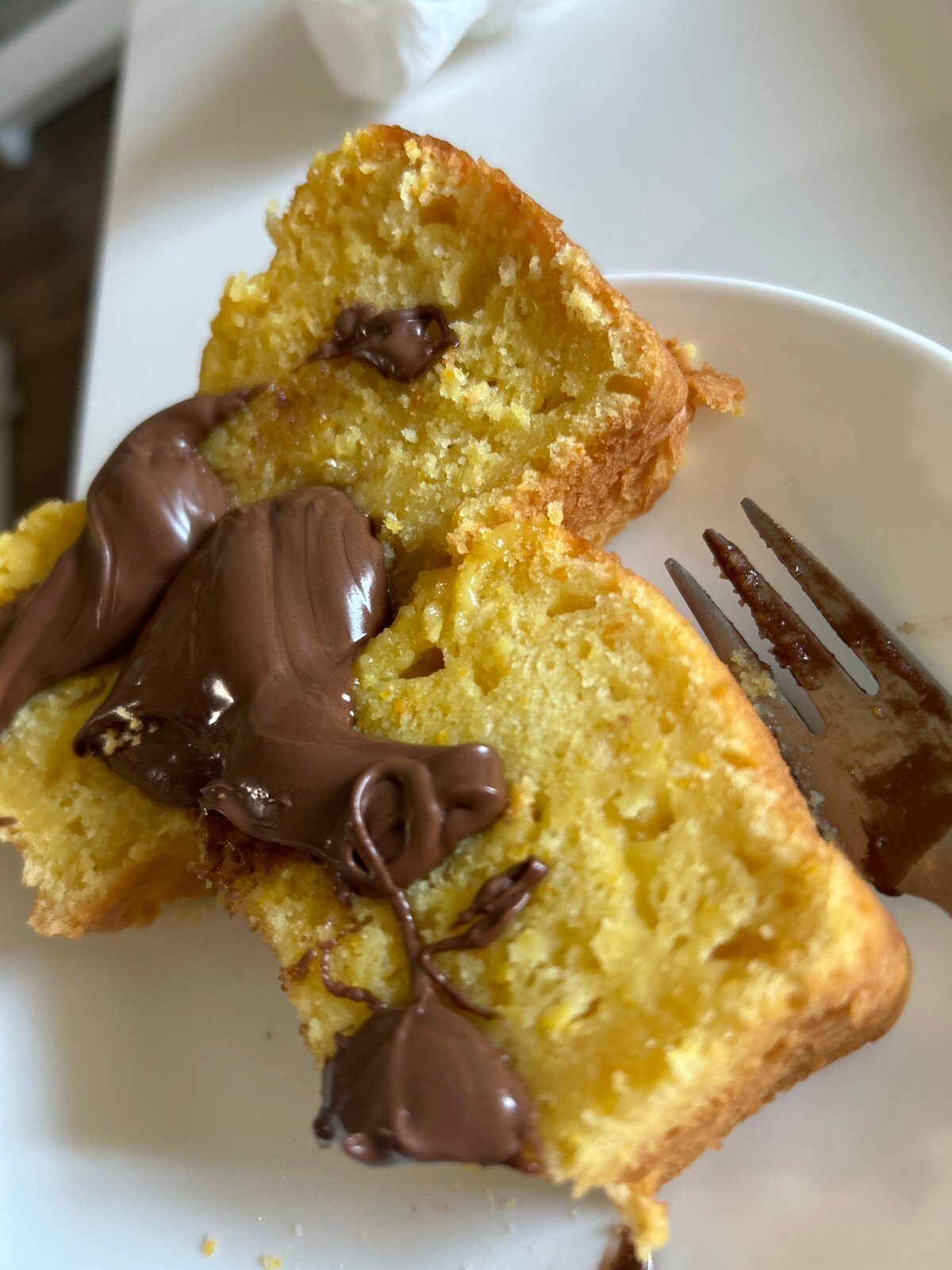 Two slices of yellow Clementine Cake topped with melted chocolate spread are on a white plate, next to a fork with some chocolate on its tines.