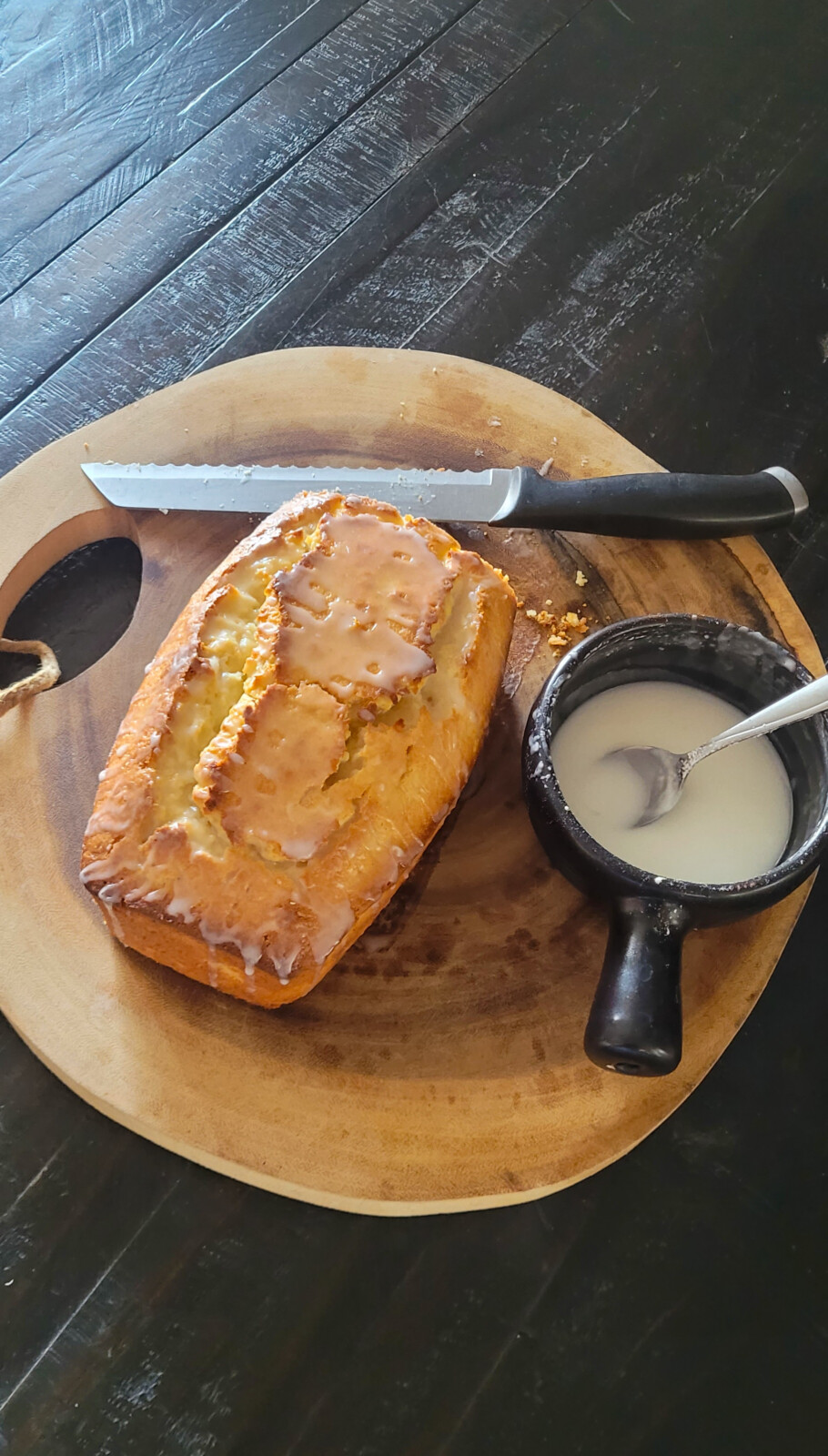 A Whole Lemon Blender Cake loaf sits on a round wooden cutting board next to a black-handled knife and a small black bowl of icing with a spoon, all placed on a dark tabletop.