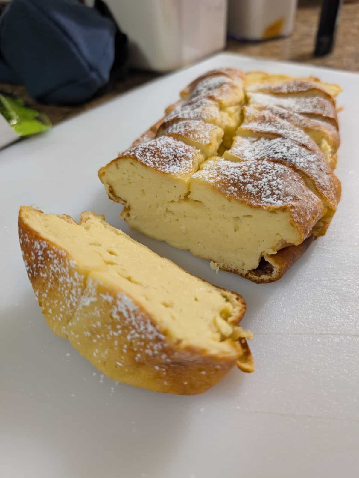 A loaf of custard-like 3 Ingredient Yogurt Cake dusted with powdered sugar, with one thick slice cut and placed in the foreground on a white cutting board.