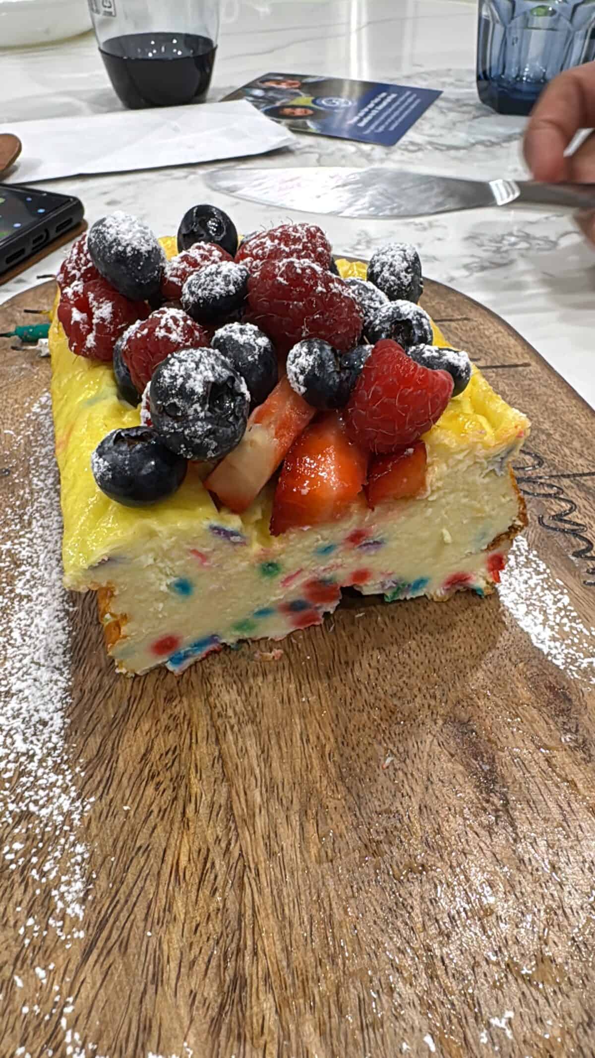Rectangular 3 Ingredient Yogurt Cake with colorful candy pieces inside, topped with powdered sugar, strawberries, raspberries, and blueberries sits on a wooden board. A hand holds a fork nearby as other table items appear in the background.