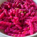 A white bowl filled with vibrant Barbie Pasta, its bright pink hue complemented by small pieces of chopped green herbs, sits on a light-colored wooden surface.