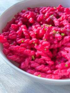 A white bowl filled with vibrant Barbie Pasta, its bright pink hue garnished with chopped green herbs, sits on a light-colored surface.