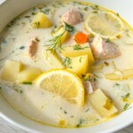 A bowl of creamy Finnish Salmon Soup with chunks of salmon, potatoes, carrot slices, lemon wedges, and fresh dill, served in a white bowl on a light wooden surface.