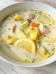 A bowl of creamy Finnish Salmon Soup with chunks of salmon, potatoes, carrot slices, lemon wedges, and fresh dill, served in a white bowl on a light wooden surface.