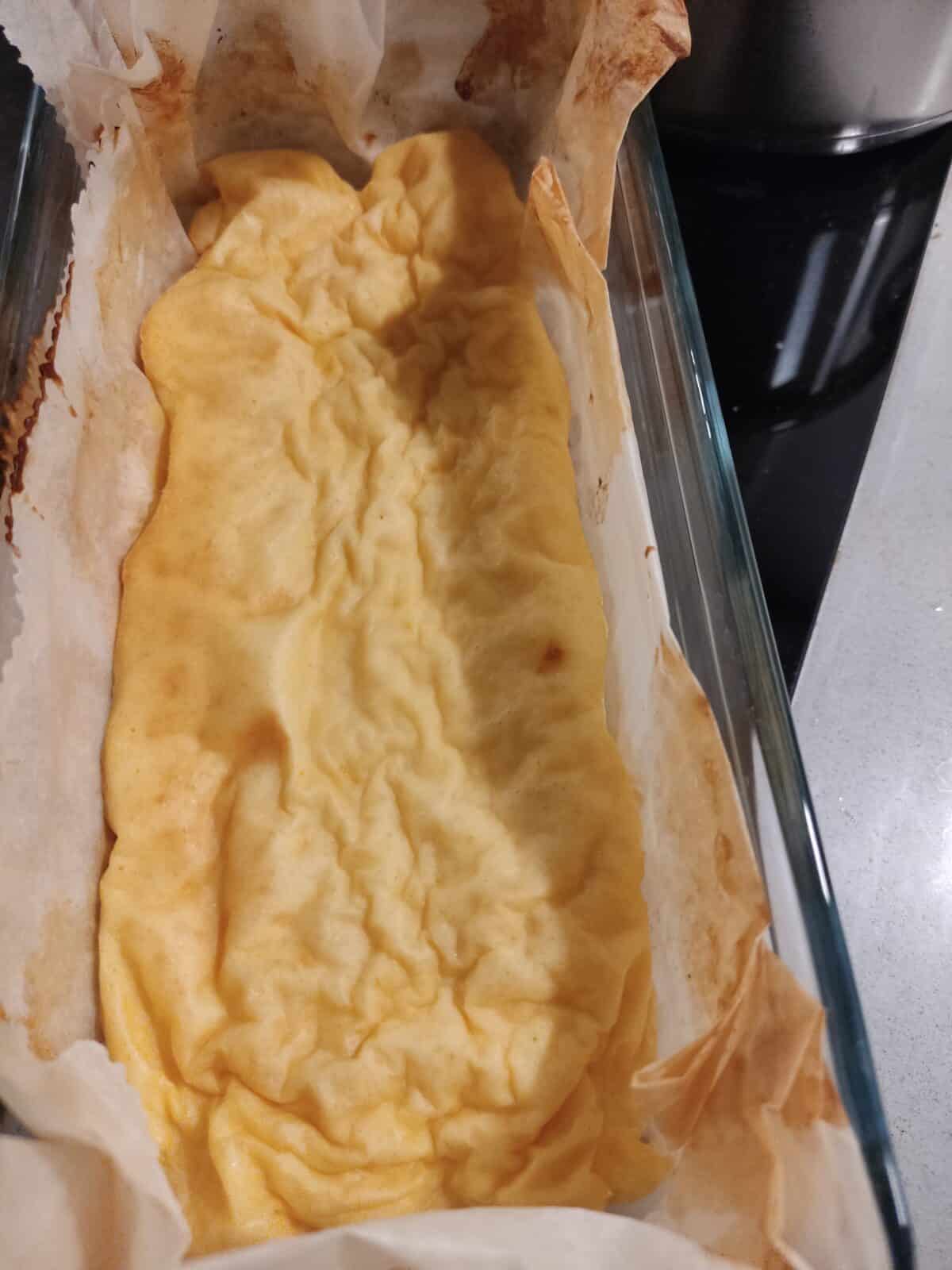 A rectangular baked dish lined with parchment paper, containing a yellow, slightly wrinkled, and puffy 3 Ingredient Yogurt Cake, possibly resembling a soufflé or baked custard, rests on a kitchen countertop.