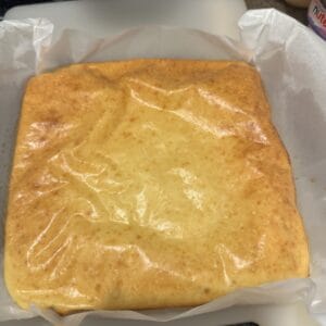 A freshly baked, golden-brown cake rests in a square baking pan lined with parchment paper, sitting on a white cutting board. The cake's surface appears smooth and slightly glossy.