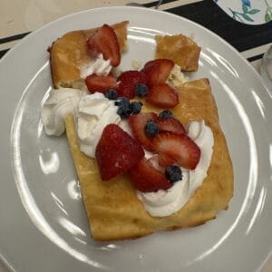A rectangular pastry topped with whipped cream, sliced strawberries, and blueberries sits on a white plate on a patterned tablecloth.