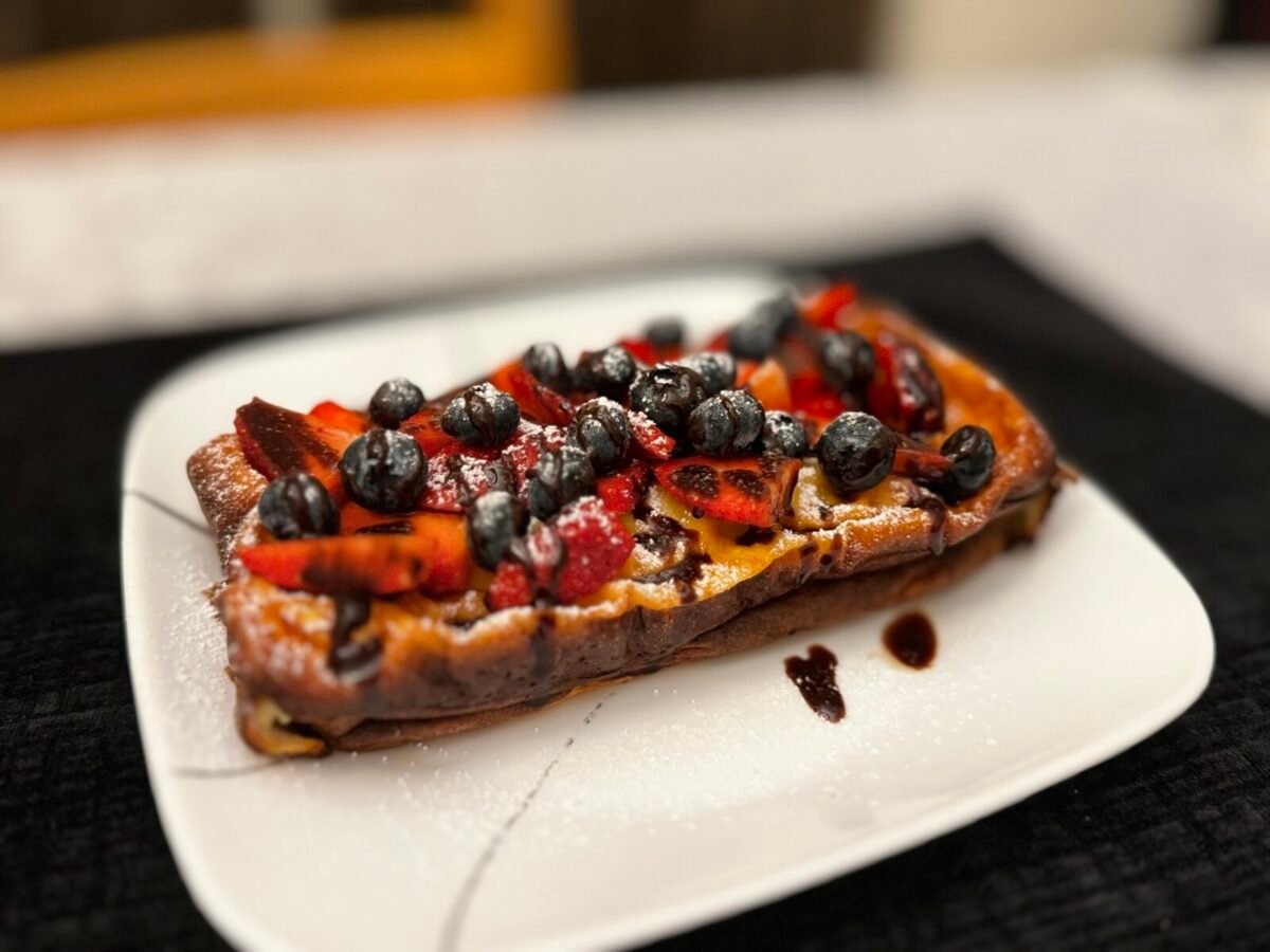 A slice of French toast topped with fresh blueberries and sliced strawberries, drizzled with chocolate sauce and sprinkled with powdered sugar, served on a white plate—perfect alongside a light 3 Ingredient Yogurt Cake.