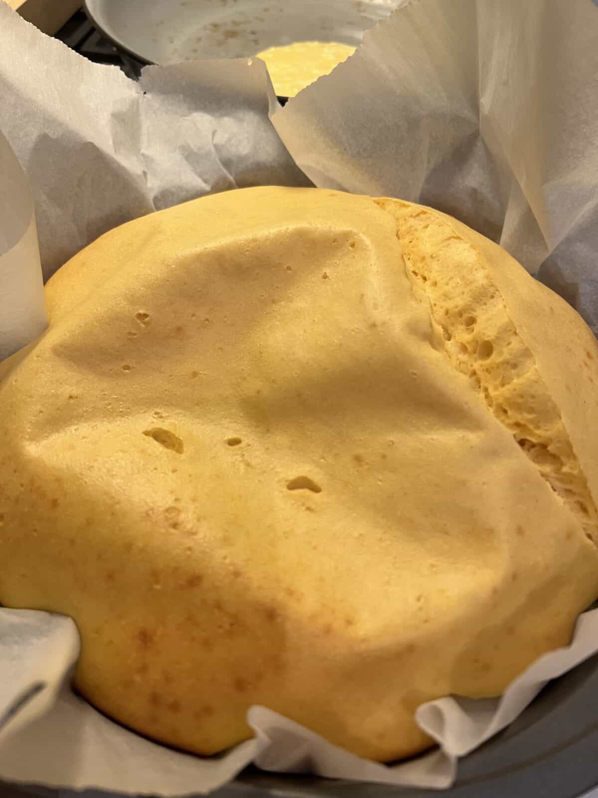 A close-up of a freshly baked, golden-yellow 3 Ingredient Yogurt Cake with a cracked top, still in a round pan lined with parchment paper. The cake looks fluffy and slightly uneven on the surface.