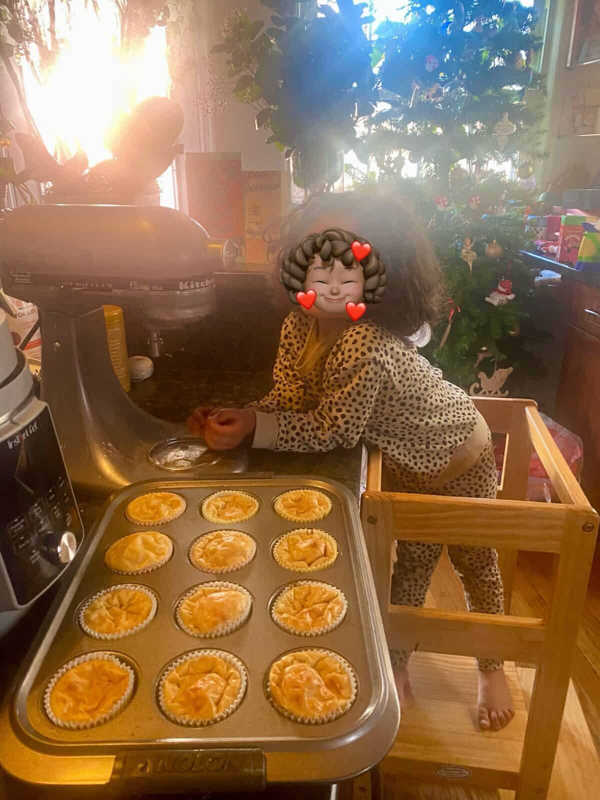 A child wearing polka dot pajamas stands on a wooden step stool in a kitchen, helping make pies. A tray of pies is on the counter. A Christmas tree is decorated in the background. The child's face is covered with a heart-eyed emoji.