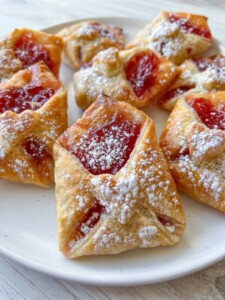 A plate of golden-brown Love Letter Pastries filled with bright red fruit jam, dusted with powdered sugar, arranged beautifully on a white plate.