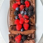 A Chocolate Yogurt Cake loaf topped with fresh raspberries, blackberries, and blueberries is served on a white oval plate. Two slices are cut from the loaf, revealing its rich, moist texture.