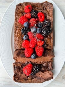 A Chocolate Yogurt Cake loaf topped with fresh raspberries, blackberries, and blueberries is served on a white oval plate. Two slices are cut from the loaf, revealing its rich, moist texture.