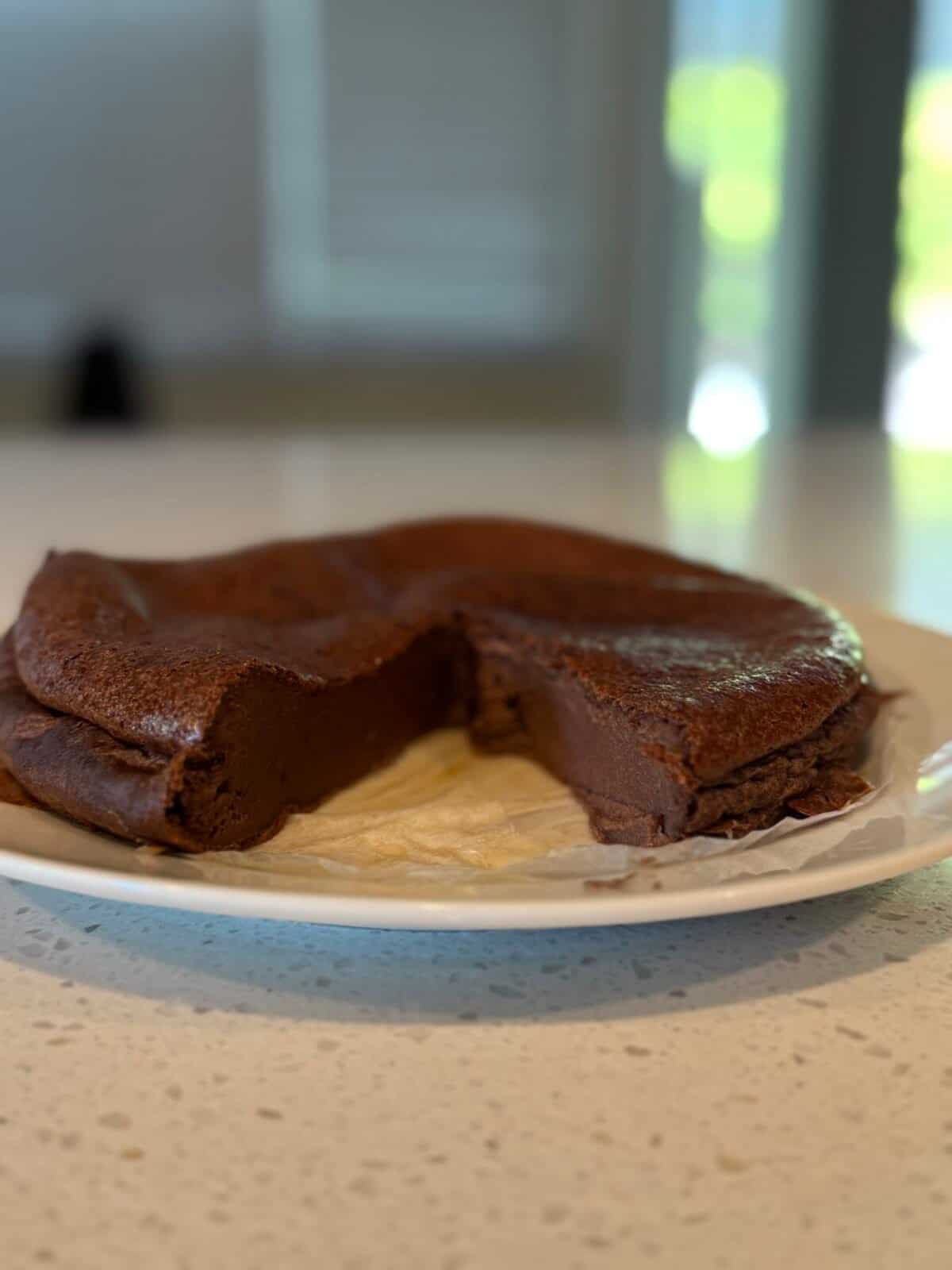 A rich, dense chocolate cake with a slice missing sits on a white plate atop a light-colored countertop, with a blurred background.