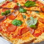 A close-up of a Low Carb Pizza topped with pepperoni and fresh basil leaves, sliced into pieces and served on a white plate. The melted cheese and tomato sauce are visible in bright, natural sunlight.