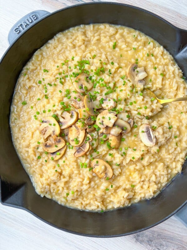 A black Staub pan filled with creamy mushroom risotto, made using a simple risotto recipe, is garnished with sautéed mushroom slices and chopped chives, and sits on a light wooden surface.