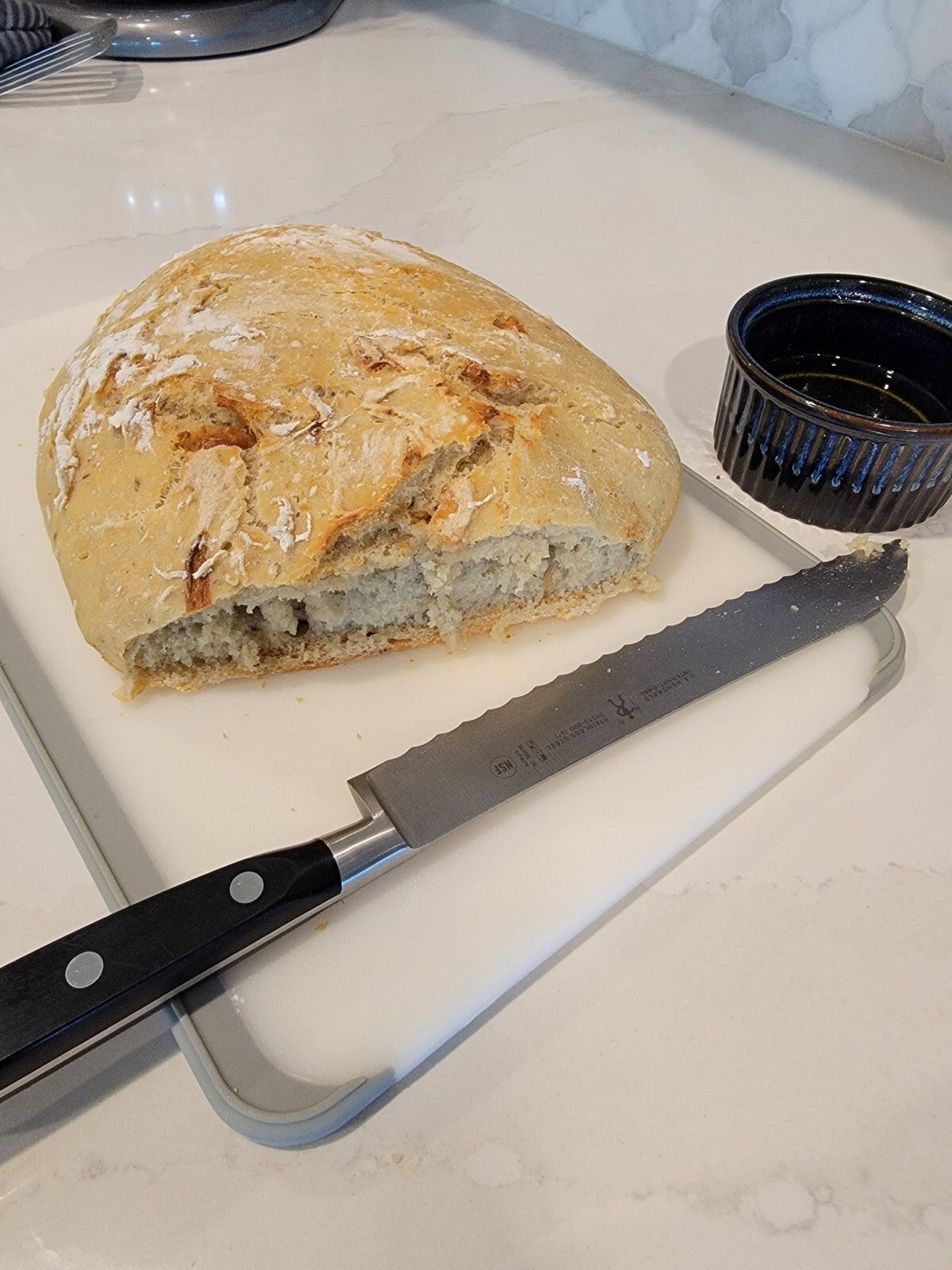 A partially sliced loaf of rustic No Knead Garlic Bread sits on a cutting board next to a black-handled bread knife. A small black bowl with olive oil is on the right, all on a white countertop.