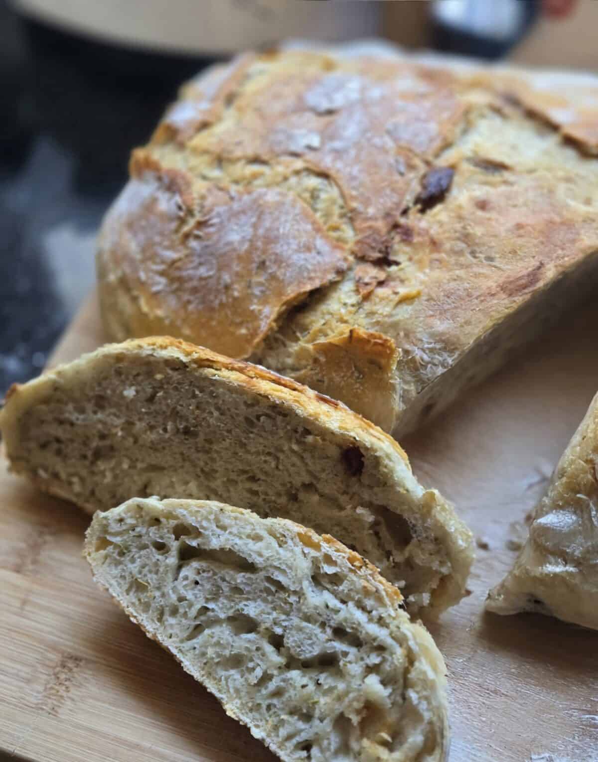 A close-up of a rustic No Knead Garlic Bread loaf on a wooden cutting board, with two slices cut to reveal the airy, textured interior and golden-brown crust.