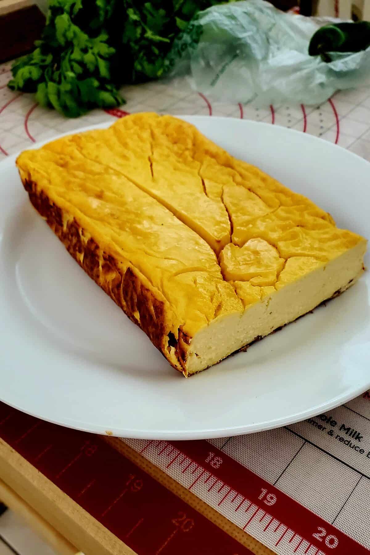 A rectangular block of golden-brown Lemon Cottage Cheese Cake sits on a white plate, with visible cracks on the surface. Fresh herbs and kitchen items are in the blurred background.
