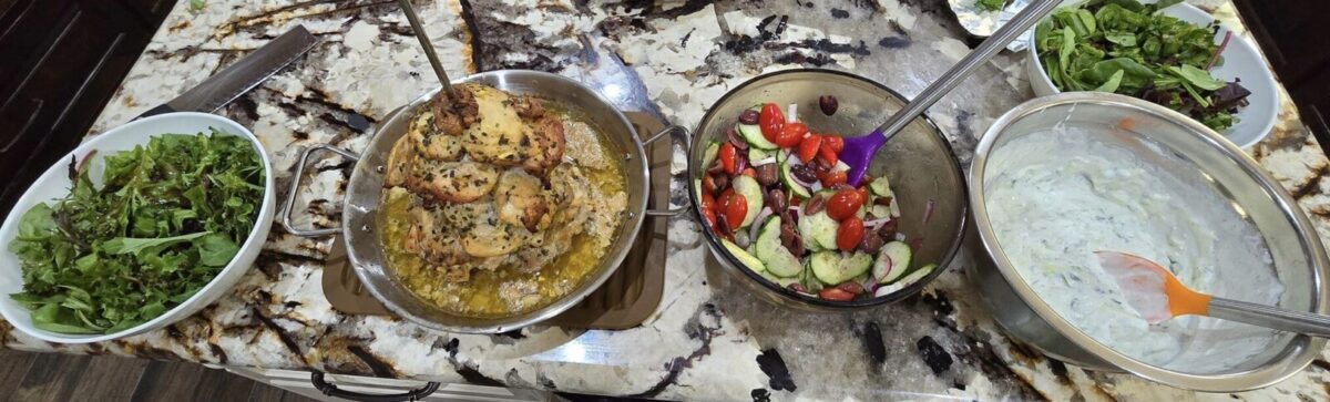 A countertop displays bowls of mixed green salad, Greek Chicken Gyros with herbs in a pan, cucumber and tomato salad, and a bowl of creamy tzatziki sauce, all ready to be served.
