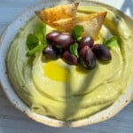 A bowl of creamy Avocado Hummus topped with black olives, fresh mint leaves, a drizzle of olive oil, and two seasoned tortilla chips sits on a light wooden surface in natural sunlight.