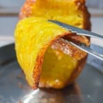 A pair of tongs lifts a crispy, golden-brown slice of Brazilian Grilled Pineapple from a larger piece on a skewer over a stainless steel tray.